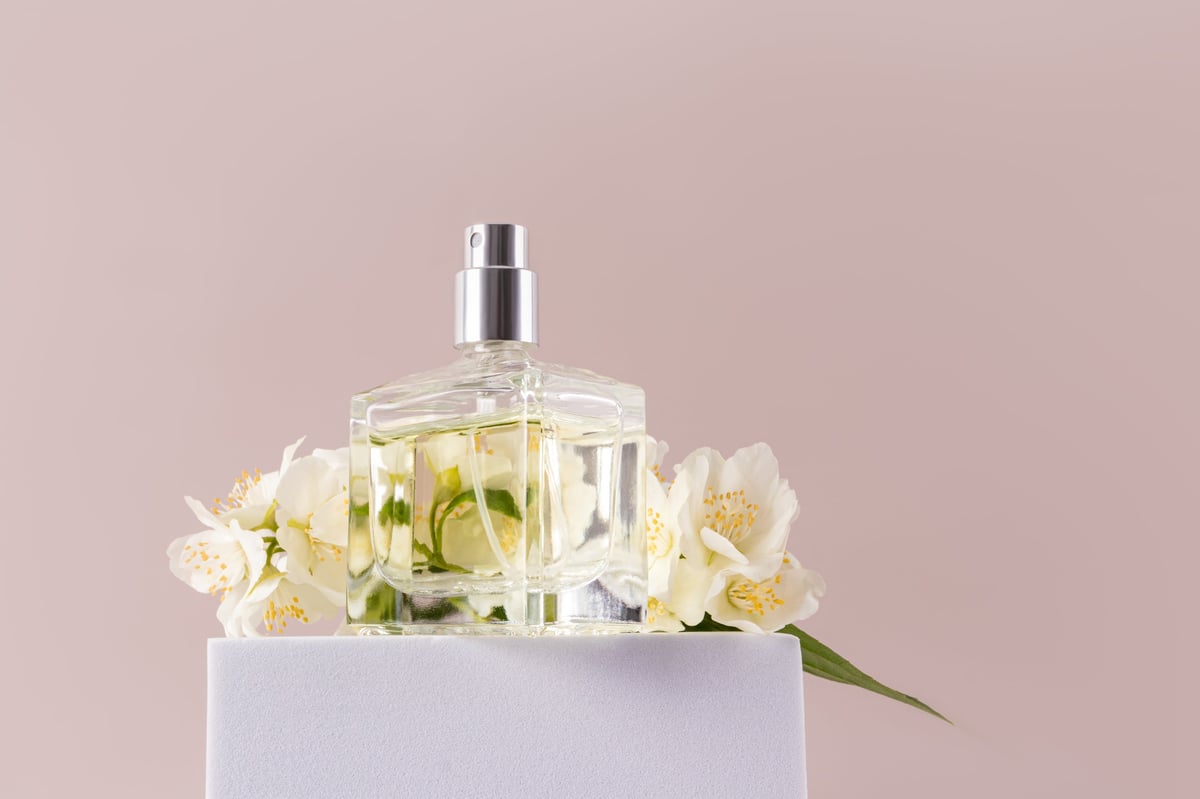 A glass transparent perfume bottle stands on a white high podium with delicate jasmine flowers. Front view. Beige background. Fragrance presentation.
