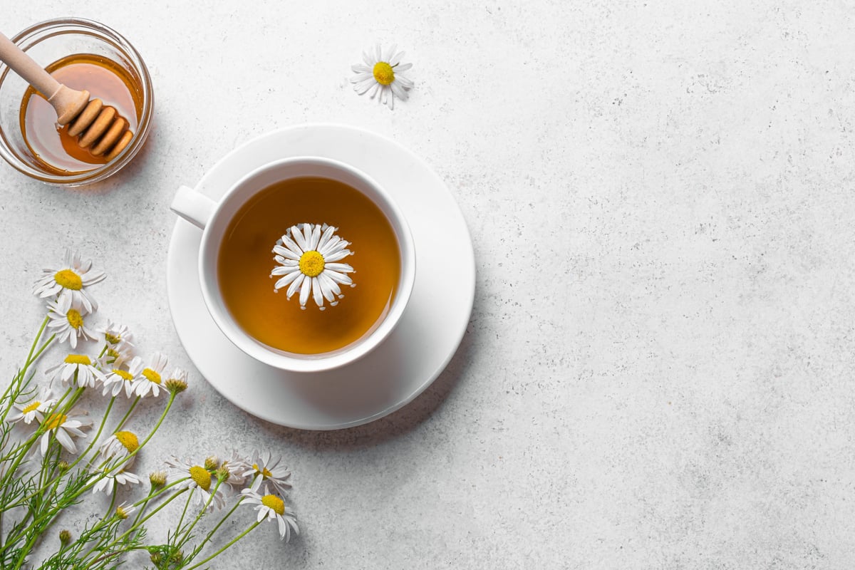 Chamomile tea cup with chamomile flowers and honey on white background, top view, copy space. Cup of calming healthy herbal tea with daisy, minimal template.