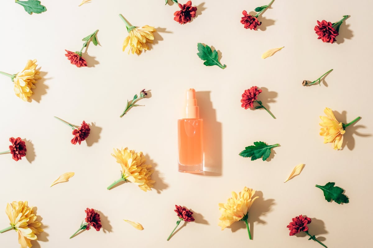 Glass cosmetic bottle with yellow and red chrysanthemums on beige background with shadows. Flat lay, natural beauty concept.