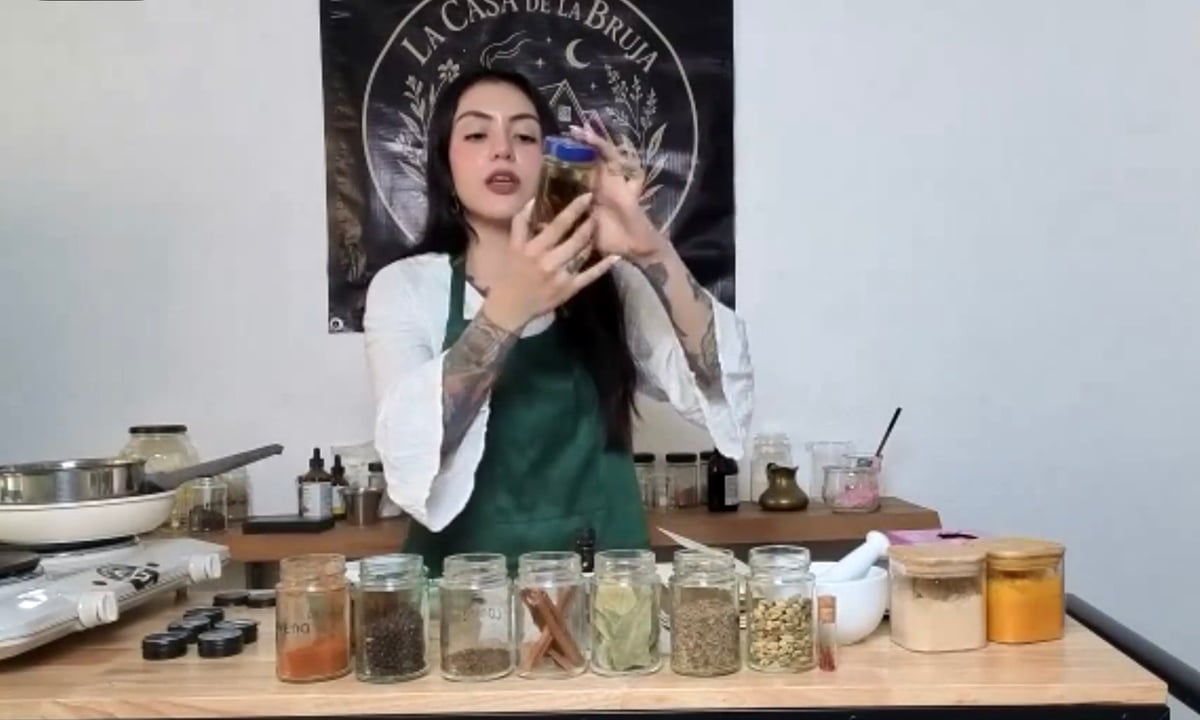 Woman demonstrating cosmetics or skincare product while seated at a table with various jars and ingredients in front of her